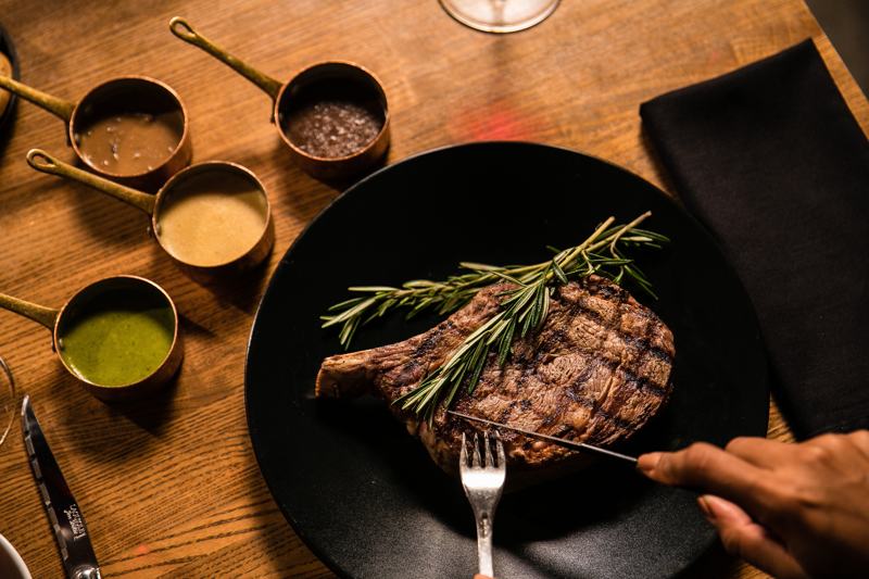 well-done-steak-on-black-plate-being-cut-with-silver-knife-and-fork-next-to-dipping-sauces